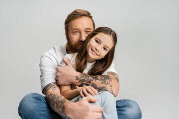 Tattooed man in t-shirt and jeans hugging happy preteen kid isolated on grey.
