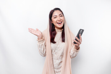 Excited Asian woman wearing hijab pointing at the copy space beside her while holding her phone, isolated by white background