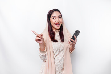 Naklejka premium Excited Asian woman wearing hijab pointing at the copy space beside her while holding her phone, isolated by white background