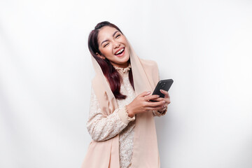 Fototapeta premium A portrait of a happy Asian Muslim woman wearing a headscarf, holding her phone, isolated by white background