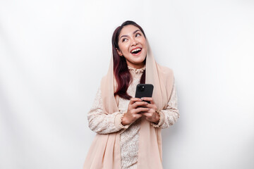 A portrait of a happy Asian Muslim woman wearing a headscarf, holding her phone, isolated by white background