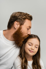 Bearded dad kissing head of smiling preteen daughter with closed eyes isolated on grey.