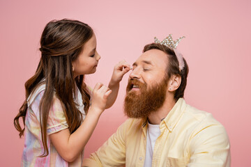 Smiling preteen girl applying eyeshadow on bearded dad with crown headband isolated on pink.