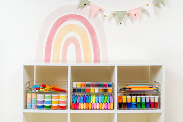 Transparent plastic containers with stationery and supplies for drawing and craft on shelves. White shelving with various material for creativity and art activity. Organizing and storage craft room.
