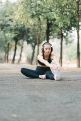 A beautiful sportswoman in sportswear is stretching her body with a smile on her face, while doing flexibility exercises to warm up before a running workout in Autumn city park background.