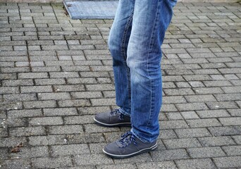 Alter schlanker Mann mit grauen Turnschuhen und blauen Jeanshosenbeinen steht auf grauem...