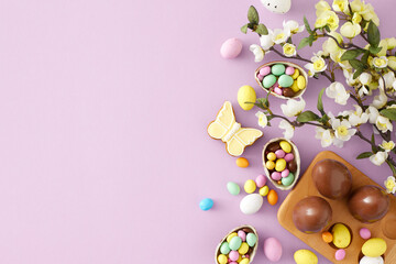 Easter concept. Flat lay photo of chocolate eggs in wooden holder dragees butterfly shaped gingerbread and spring blossom flowers on isolated violet background with copyspace. Holiday card idea