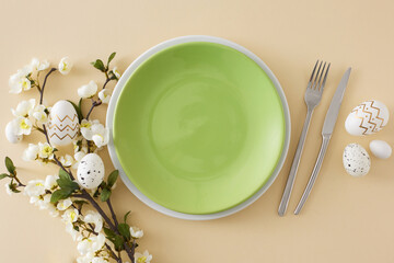 Easter decor idea. Flat lay photo of empty green plate white golden easter eggs cutlery fork knife spring blossom branch on isolated beige background