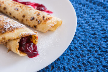 Homemade pancakes with preserved cherry jam, sprinkled with powdered sugar. Blue crochet napkin under the plate. 
