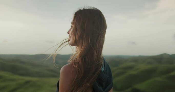 Woman enjoy green hills landscape at sunset. Long hair brunette girl looking on beautiful nature scene, hair blowing in wind. Freedom, happiness lifestyle. Travel, tourism, holiday. Slow motion 4k