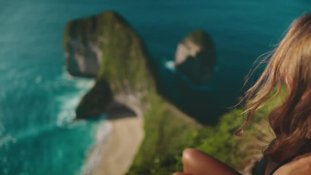 Young woman tourist sitting over Tyrannosaur rock at Kelingking Beach, Nusa Penida, Indonesia. Famous touristic place. Girl hiking backpacker traveler relaxes, sunset sea view point. Slow Motion shot