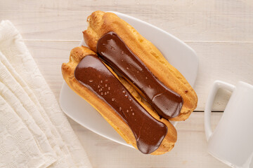 Two chocolate eclairs with white ceramic saucer and cup on wooden table, close-up, top view.