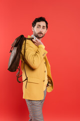 Trendy man in jacket holding brown backpack isolated on coral red.
