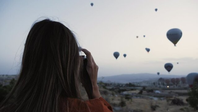 Rear view happy young Caucasian tourist woman using film camera to take picture of incredible hot air balloons sky view.