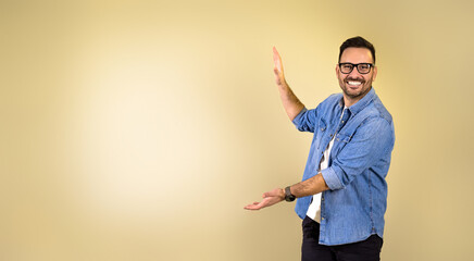 Portrait of smiling young adult man wearing blue denim shirt and eyeglasses gesturing towards copy space while standing isolated over beige yellow background