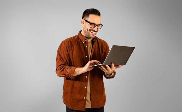 Focused smiling young adult handsome businessman dressed in shirt working over laptop computer while standing confidently on isolated gray background