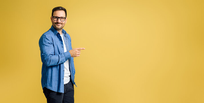 Portrait of smiling charming young adult businessman wearing denim shirt and eyeglasses pointing away at copy space isolated on yellow background