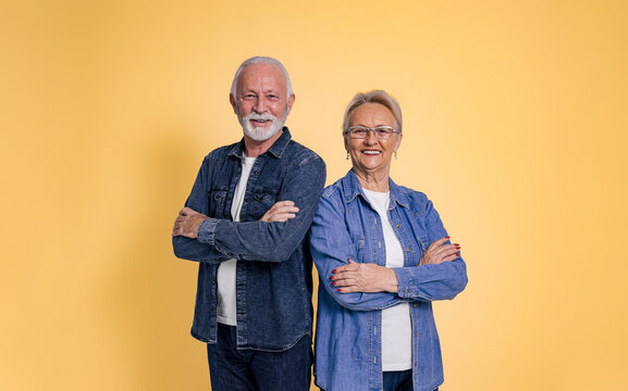 Portrait of happy and confident senior couple wearing denim shirts with arms crossed posing while standing isolated over yellow background
