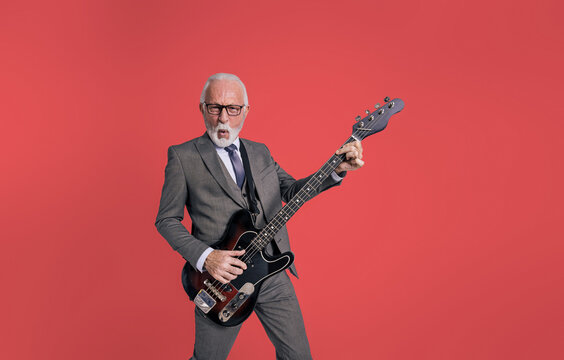 Portrait of excited bearded senior businessman dressed in elegant suit singing cheerfully and playing electric guitar isolated over red background