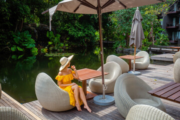 Happy travel woman on vacation in modern tropical resort. Female traveler in yellow dress and straw hat enjoy her trip, sitting in outdoor restaurant near beautiful pond at summer sunny day.