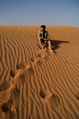 Fototapeta na wymiar Male tourist looking away while sitting on sand dunes in desert at Dubai, United Arab Emirates
