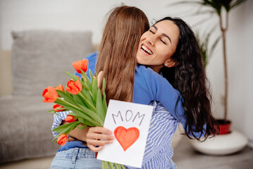 Happy mother's day. Child daughter congratulates mom and gives her flowers tulips and postcard. Mum and girl smiling and hugging. Family holiday and togetherness.