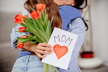 Grateful mom hugging daughter girl, holding flowers bouquet, receiving hand drawn greeting card...