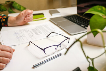 Fototapeta na wymiar Eyeglasses for office work. Office workplace. White wooden table with office supplies desktop computer and book, green plant, female hands. Out of focus