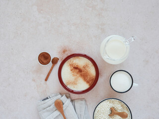 dessert of rice pudding portion served with cinnamon powder on top along with the ingredients for the preparation: milk, sugar, cinnamon, lemon and rice. Top view on a pink marbled kitchen countertop.