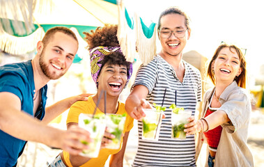 Diverse trendy friends having fun drinking mojito cocktails at sunset beach party - Summer joy and life style concept with fancy people at chiringuito happy hour - Warm filter with focus on left woman
