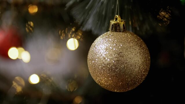 Golden Shiny Christmas Ball Hanging on a Spruce Branch on a Background of Lights. Close up. Christmas tree decoration bauble ball. Blurred background with blinking LED lights. Christmas decoration.