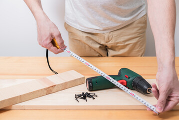 A man works with an electric screwdriver on a wooden table, also makes measurements with a tape...