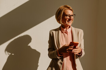 Senior businesswoman using smartphone while standing over a white wall in an office