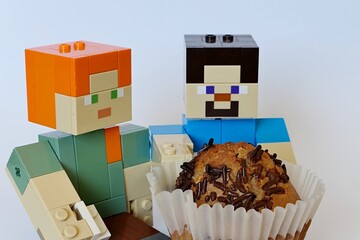 Fototapeta premium LEGO Minecraft large figures of Steve and Alex, Steve is holding huge muffin cake with chocolate chips on top in a white paper cup. Light grey background.