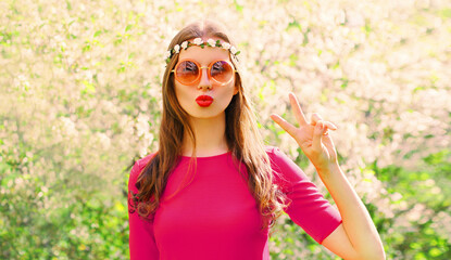 Portrait of beautiful hippie young woman blowing her lips sending air kiss wearing floral headband, sunglasses in spring blooming garden on flowers background - Powered by Adobe