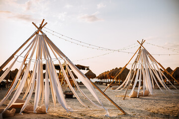 Boho tents made of wooden bars and white material on the beach. In the background, close to the...