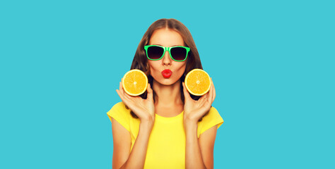 Summer portrait of stylish young woman blowing her lips sends kiss with slices of fresh orange fruits wearing sunglasses on blue background