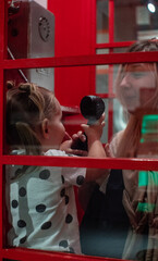 In a traditional red telephone booth, mother and baby are holding a phone in their hands and talking. Mom shows a small child how to speak in a telephone booth on the phone.