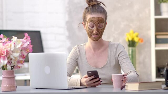 Woman cosmetic facial skin care. Woman in skin rubber peeling face beauty mask sitting at desk using phone and laptop.