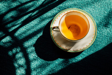 dainty white teacup with green and gold border in the afternoon sun on teal rug, carpet with shadows