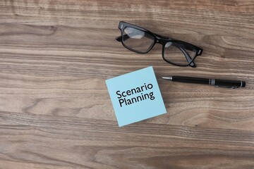 A post-it note with a message on Scenario Planning, a pen, and eyeglasses on the wooden office table. View from the top, flat lay 