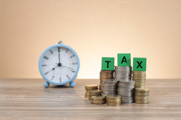 The word TAX is written on the green wooden blocks on top of the stacked coins. An alarm clock at the back 