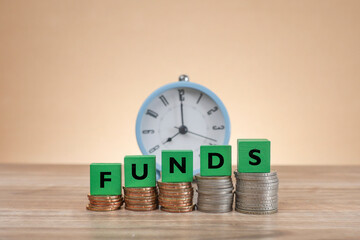 The word FUNDS is written on the green-colored wooden cubes on top of the stacked coins.  An alarm clock at the back