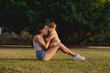 "Young mother enjoying happy moment with baby sitting on sunny park grass"

