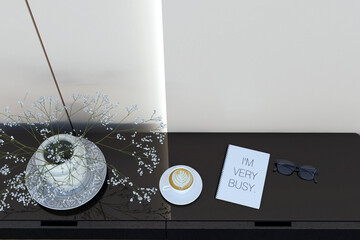 Top view of long black table next to a white wall with a vase of white roses, coffee cup, lens and a notebook that says "i'm very busy".