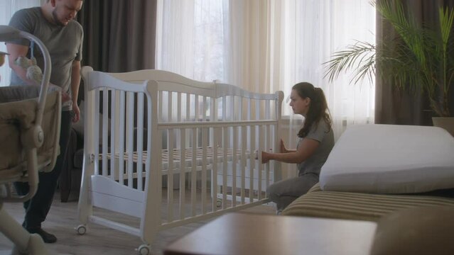 Loving parents together assembling white crib for future baby at nursery. Young father uses hex key for fixing mounts. Caring wife helps beloved man. Concept of parenthood, childhood and family.