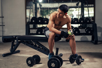 Fitness man shirtless sitting drinking the water after weight training in fitness gym. Health care and workout. Asian man replenishing water balance after workout. Concept of health and wellness.