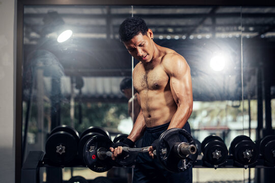 Asian man bodybuilder shirtless effort weight training biceps curl with barbell in fitness gym. Weight training exercise in concept of health and wellness.