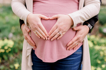 Maternity photo, pregnant woman has her hand in a heart shape over her belly, her husband's hands are also on her pregnancy bump.