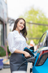 Woman plugging in the charger into a socket of her blue electric car at a charging station in the street